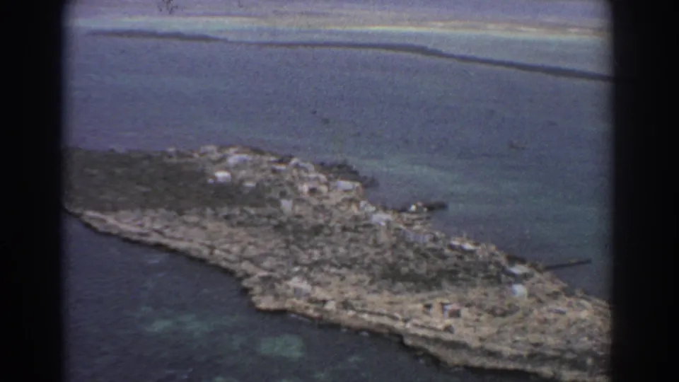 Late-frame 1967 Perth-area aerial view showing the island shoreline, scattered structures, reef-toned water, and visible 8mm film grain.