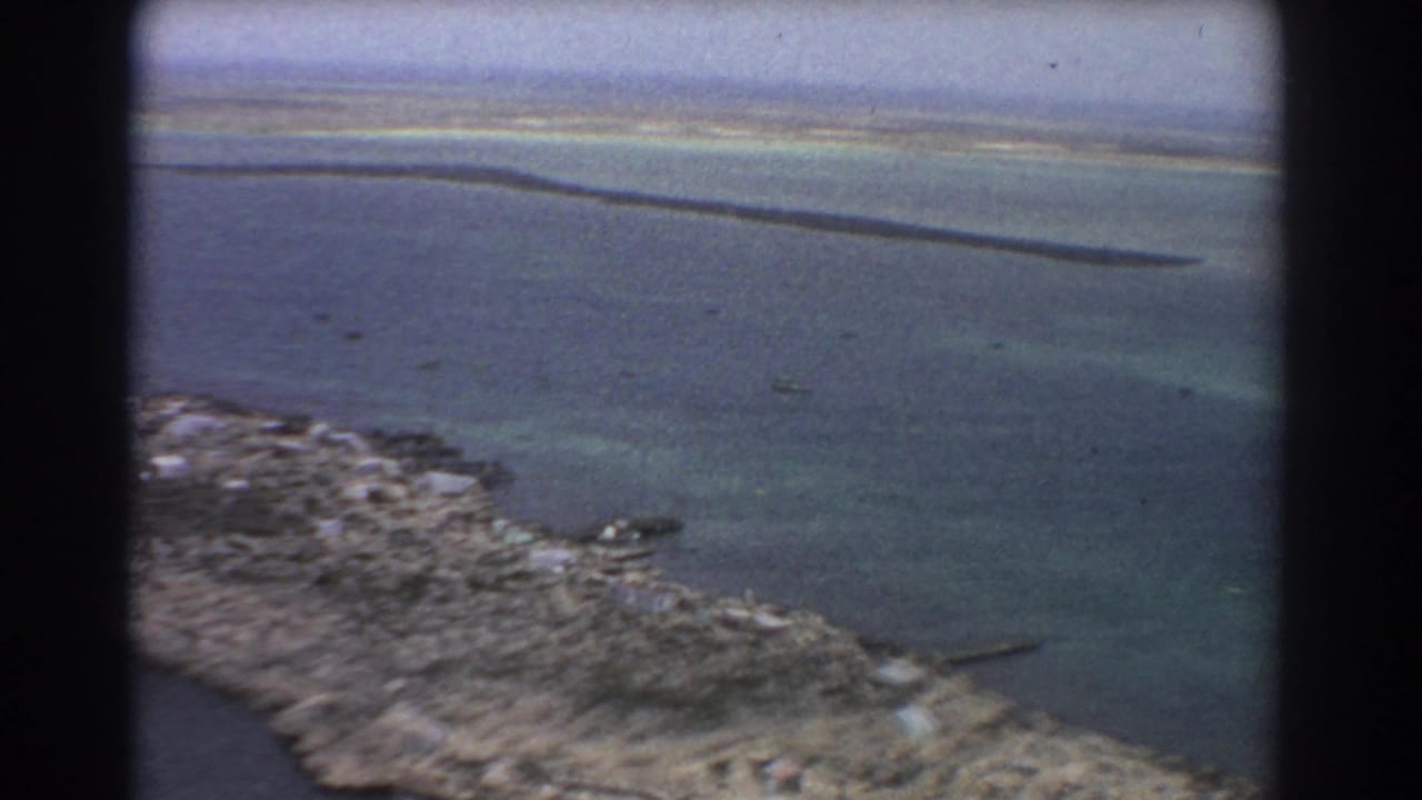 1967 8mm aerial view near Perth showing blue-green water, rocky edge, shallow patches, and narrow sandbars under hazy light.