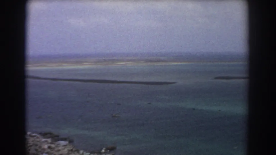1967 8mm aerial frame near Perth with calm blue water, pale shallows, narrow sandbars, and a hazy distant horizon.