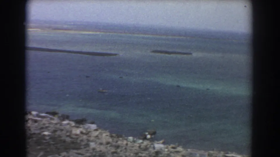 1967 8mm aerial frame near Perth showing turquoise shallows, darker water, rocky lower edge, and elongated sandbars.