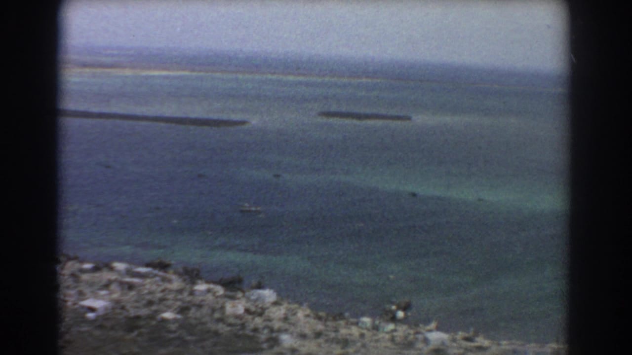 Aerial view over blue-green ocean with rocky shoreline at lower left and distant narrow landforms near the horizon.
