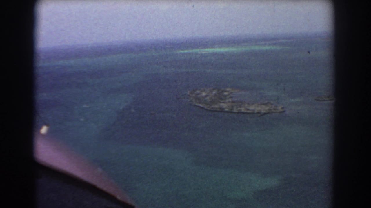 1967 8mm aerial footage near Perth showing a small offshore island surrounded by dark blue and turquoise water through an aircraft window.