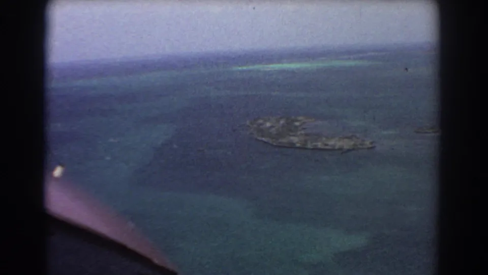 1967 8mm aerial footage near Perth showing a small offshore island surrounded by dark blue and turquoise water through an aircraft window.