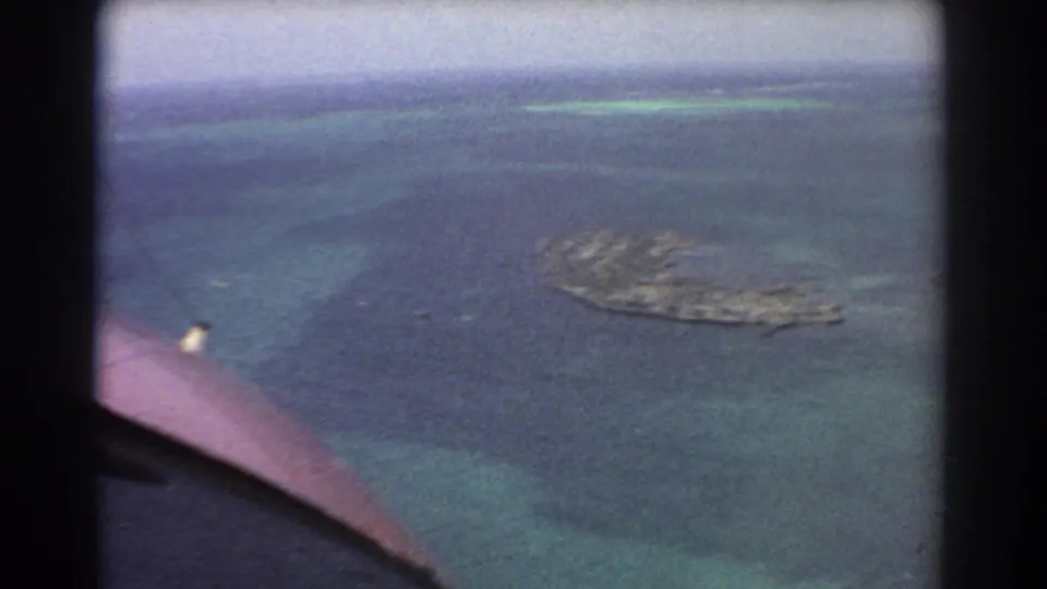 A 1967 home-movie aerial view near Perth shows an offshore island, shallow reef-colored water, and the edge of the aircraft window in frame.