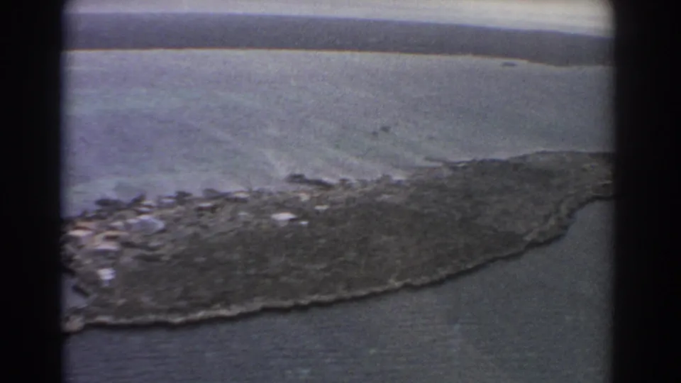 1967 8mm aerial view near Perth showing a rocky coastal point, calm ocean, a small boat offshore, and a low shoreline in the distance.