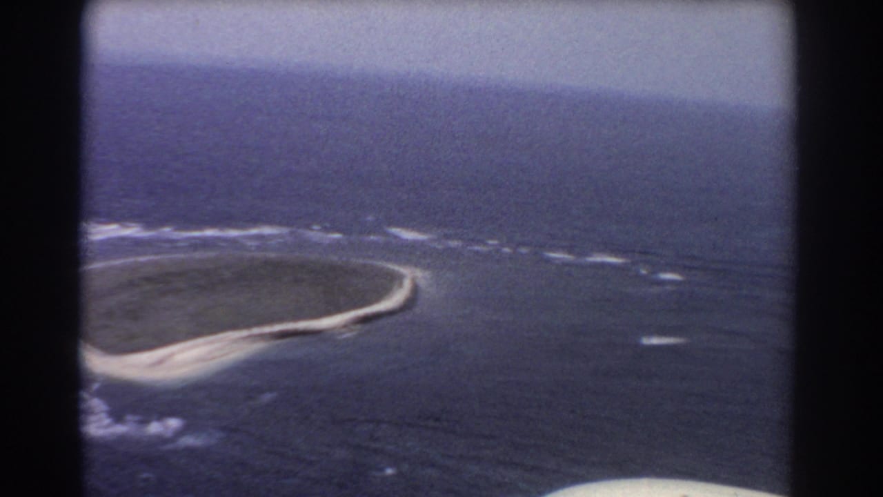 Aerial 8mm frame of a small atoll-like reef and sandbar surrounded by ocean, with white surf breaking along the edge.