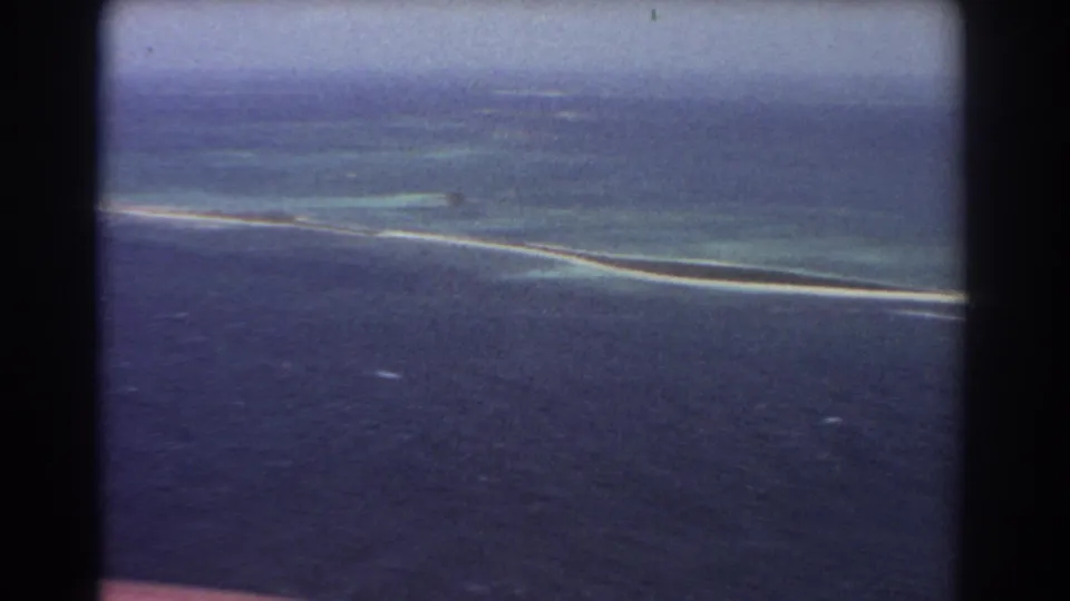 Aerial 1967 8mm frame showing a long sandbar with turquoise shallows and darker surrounding ocean under a soft vintage color shift.