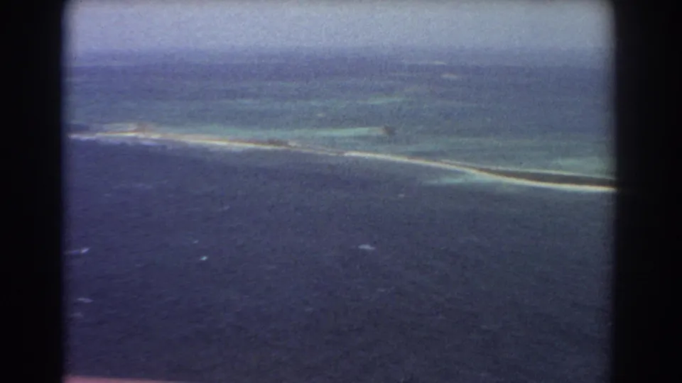 1967 8mm aerial view near Perth, Australia, showing deep blue water, turquoise shallows, and a long pale sandbar or reef below the airplane.