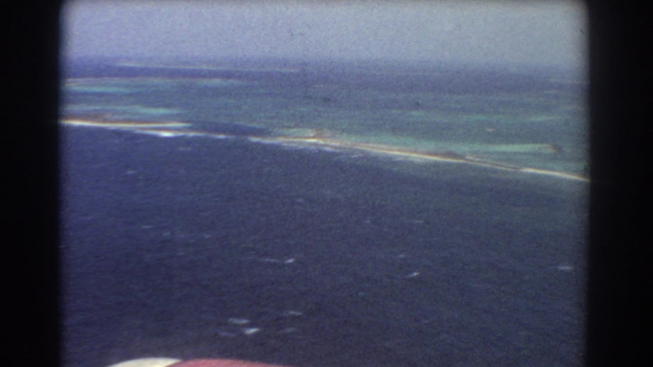 1967 8mm frame from an airplane near Perth, Australia, with dark ocean foreground, turquoise reef shallows, and a low horizon in soft focus.