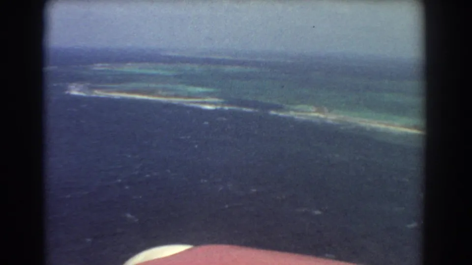 Oblique 1967 home-movie aerial view near Perth, Australia, with a curved reef or sandbar separating dark ocean from pale green shallows.