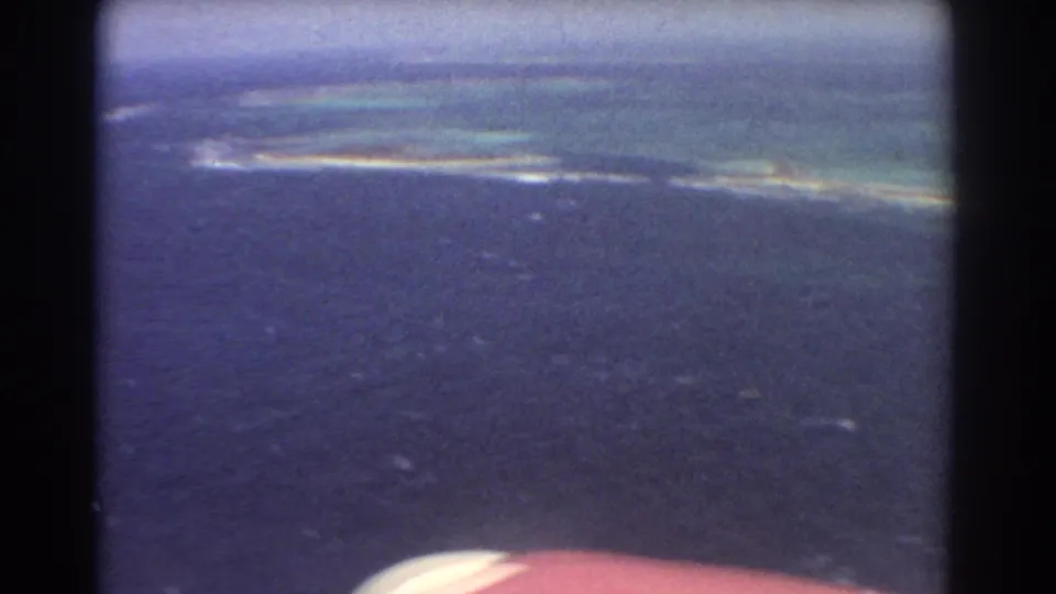 1967 8mm aerial frame near Perth, Australia, looking over dark blue ocean toward a thin bright reef line, pale shallows, and a small aircraft edge at the bottom.