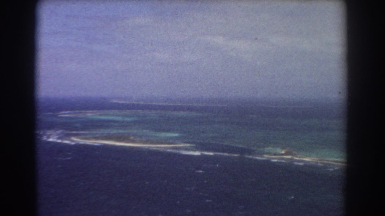 1967 8mm aerial view near Perth metadata context, showing blue ocean, reef shallows, and a thin island shoreline below a hazy sky.