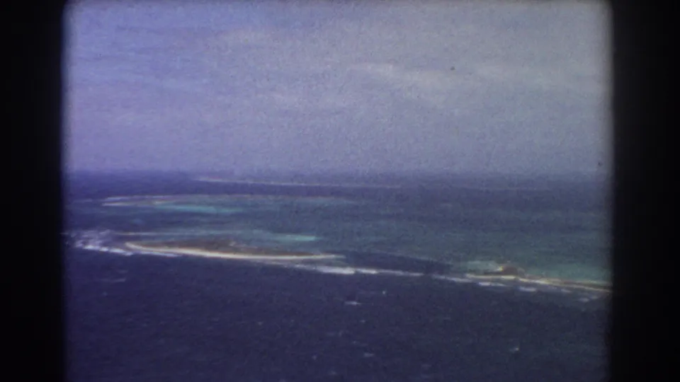 1967 8mm aerial view near Perth metadata context, showing blue ocean, reef shallows, and a thin island shoreline below a hazy sky.