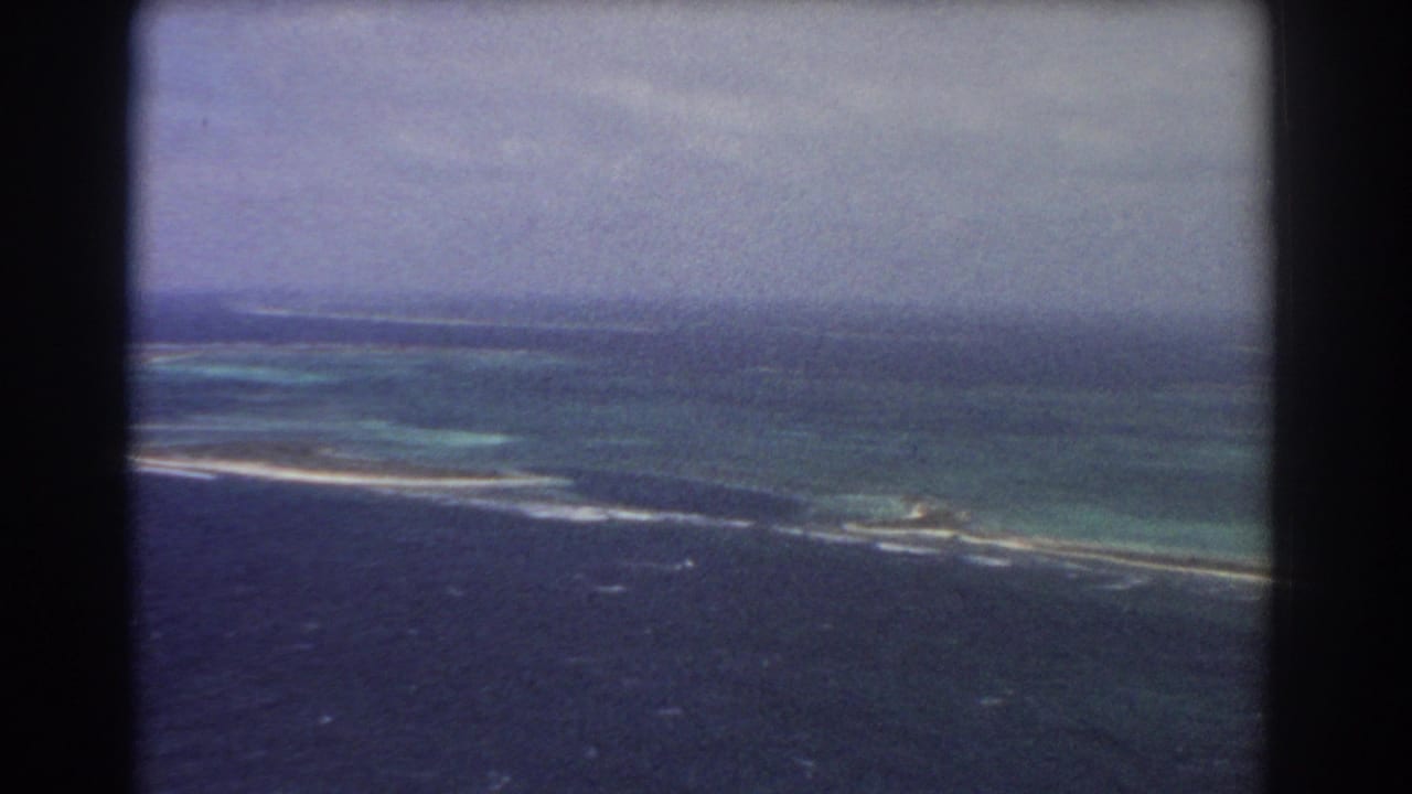 Aerial coastal view with reef patterns, white surf, distant low land, and cloudy sky.