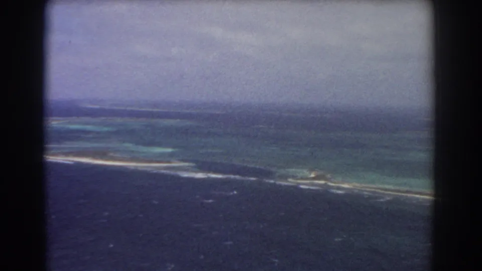 1967 home-movie aerial frame over ocean and reef near the supplied Perth location, with low coastline and white surf visible.