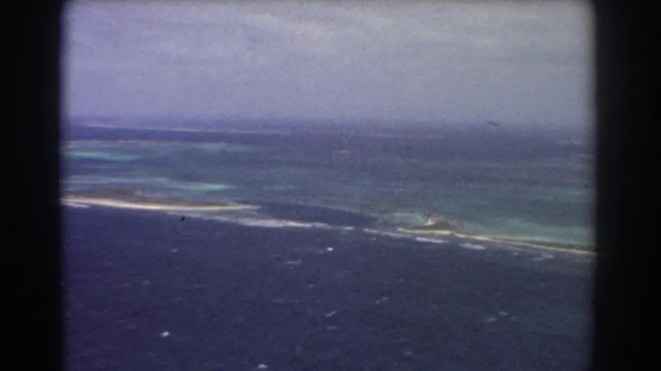 1967 8mm aerial view, supported by metadata, looking over dark ocean toward turquoise reef water and a low sandy coastline.