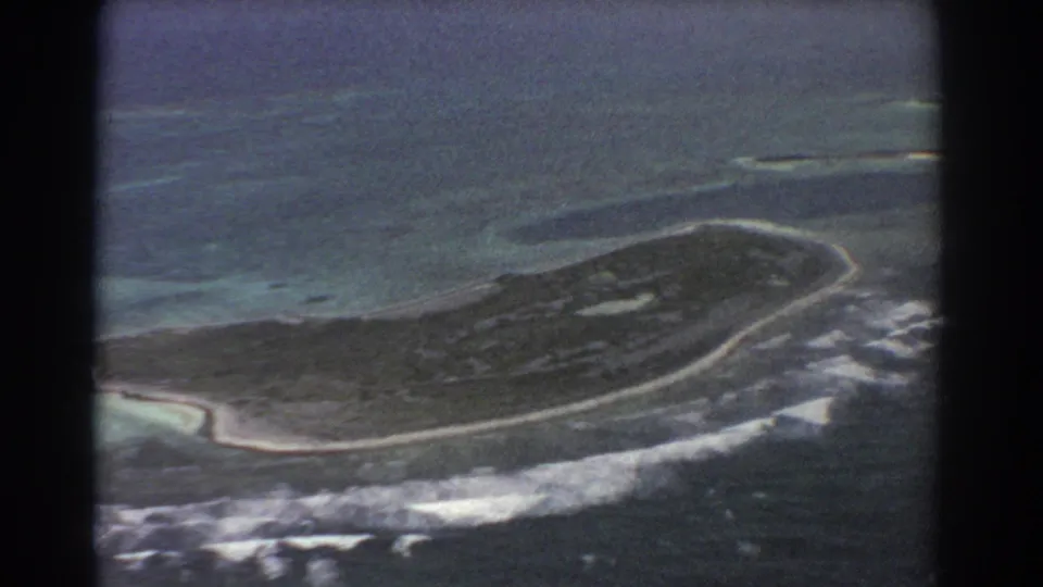 1967 8mm aerial frame of a low island or atoll-like landmass, with sandy edges, surf breaks, reef shallows, and surrounding ocean.