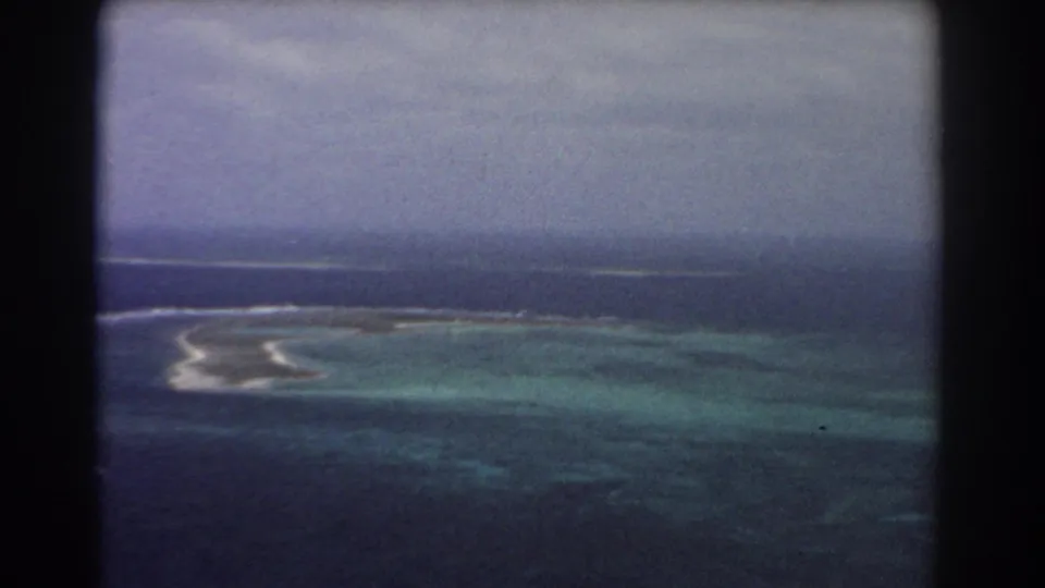 1967 8mm aerial view shows reef flats and surf bands receding toward darker sea and a cloudy horizon.