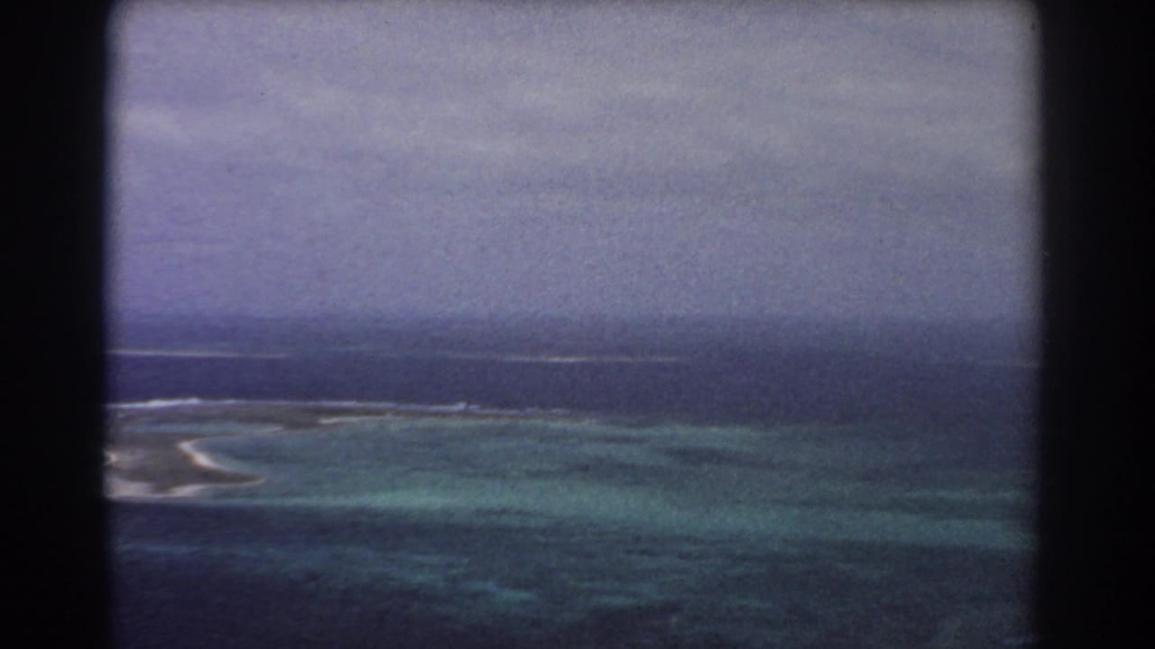 1967 8mm frame from an aircraft over reef flats and dark ocean water, with surf lines under a hazy overcast sky.