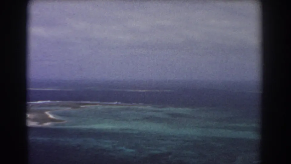 1967 8mm frame from an aircraft over reef flats and dark ocean water, with surf lines under a hazy overcast sky.