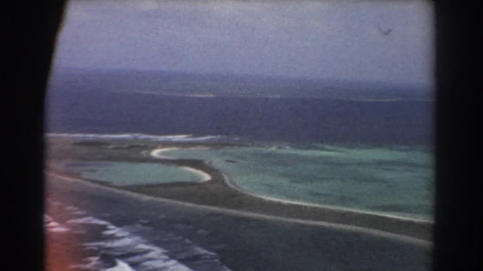 1967 8mm aerial frame showing a winding sandbar through shallow lagoon water with reef, surf, and open ocean beyond.