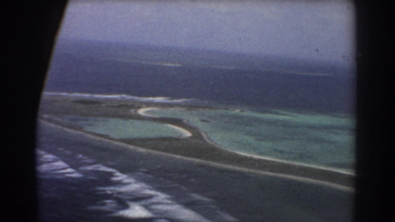 Aerial view through an aircraft window of a narrow island edge, turquoise lagoon, reef surf, and darker ocean.