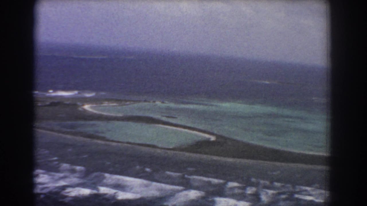 1967 aerial home-movie frame near Perth, Australia, with a curved sandbar or island edge, shallow lagoon, reef line, and dark sea beyond.