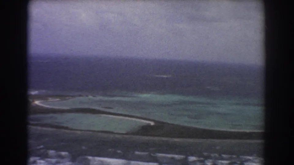 1967 8mm aircraft-window view near Perth, Australia, showing shallow lagoon color, a low island strip, reef surf, and darker offshore water.