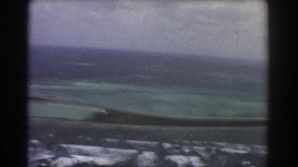 1967 8mm aerial view near Perth, Australia, with aircraft-window framing, pale lagoon shallows, a dark reef or land strip, and open ocean beyond.