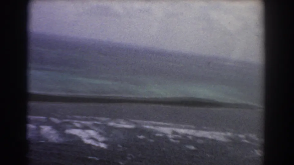 1967 Perth, Australia aerial frame with pale surf bands, blue-green water, a dark shoreline strip, and soft 8mm film grain.