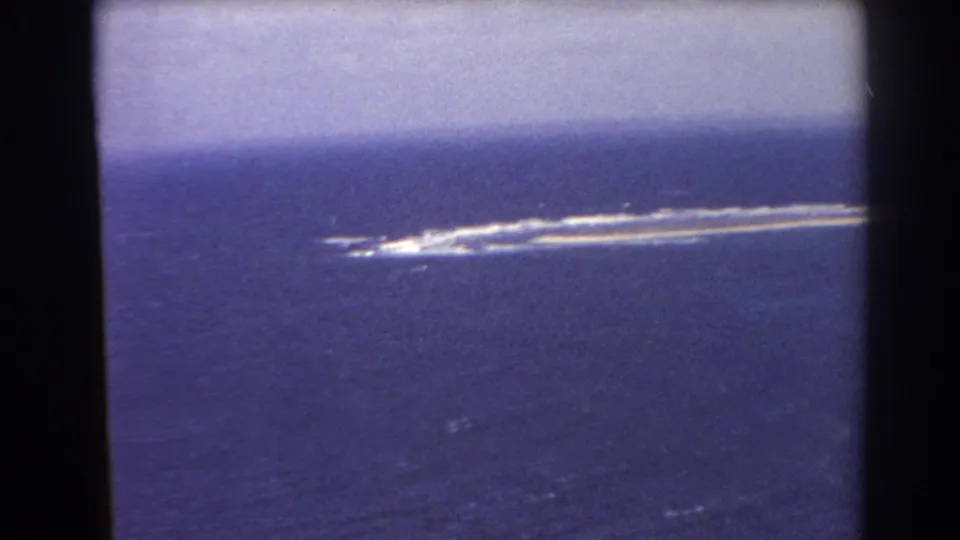 Perth-area 1967 aerial home-movie frame with a narrow pale feature, white surf breaks, blue water, and dark frame edges.