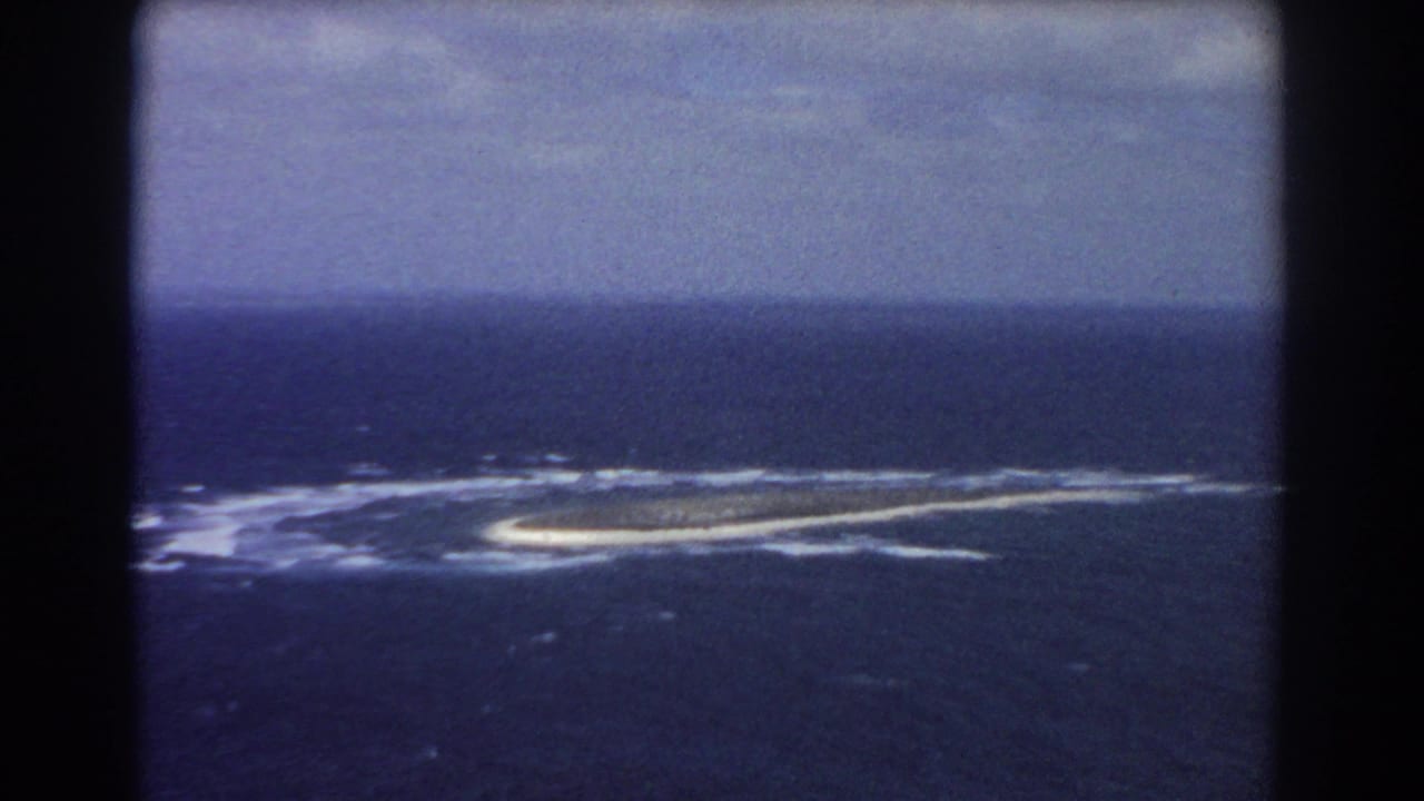 1967 home-movie aerial view identified as near Perth, Australia, showing a low island, surrounding surf, and overcast horizon.