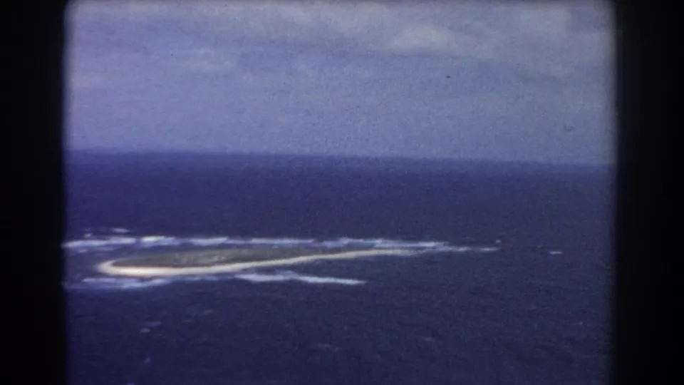Aerial 8mm frame of the identified Perth-area island, with pale shoreline, white wave breaks, and a muted cloudy sky.