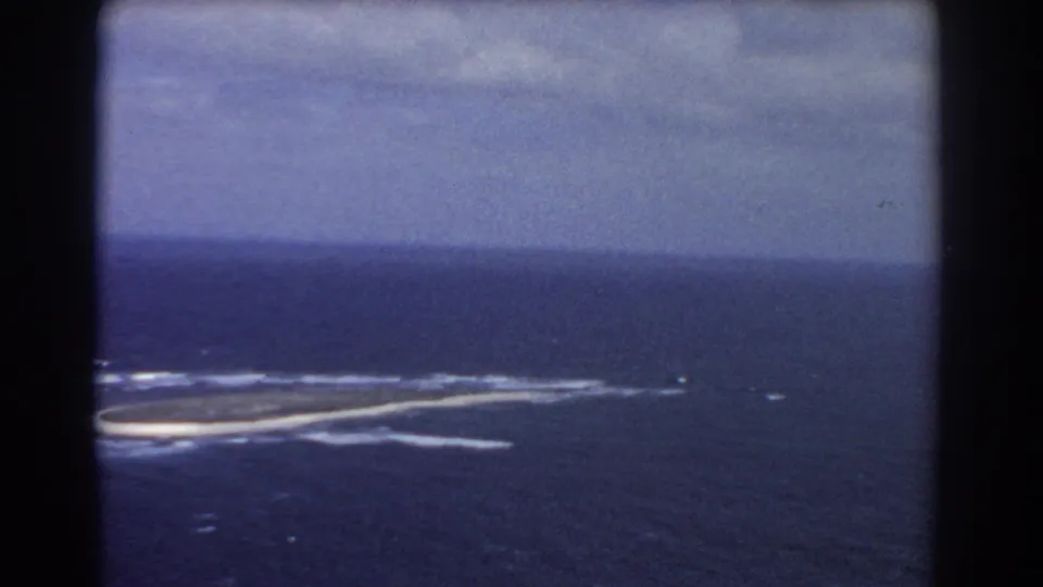 Soft, blue-tinted 1967 aerial frame showing the island partly cropped at left, white surf, dark water, and cloud cover.