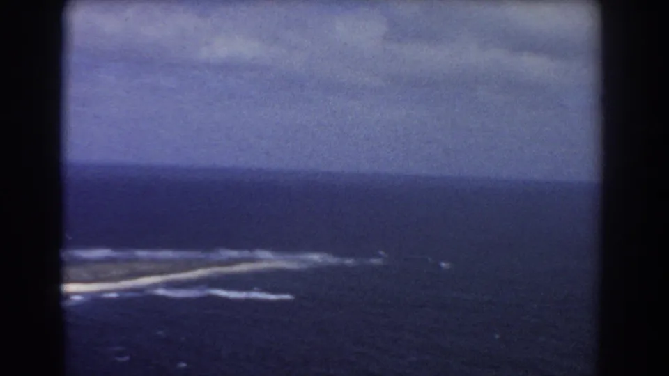 Final aerial frame from the Perth-labeled 1967 clip, with island shoreline low in frame, surf streaks, and open sea under gray clouds.
