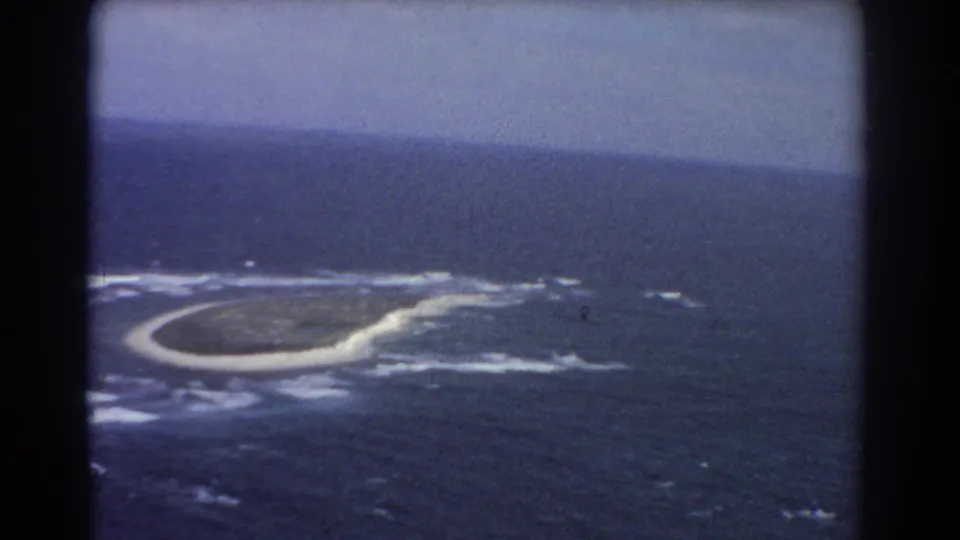 Aerial 8mm frame from near Perth showing a crescent-like island, pale sandbar, white breakers, and surrounding blue water.