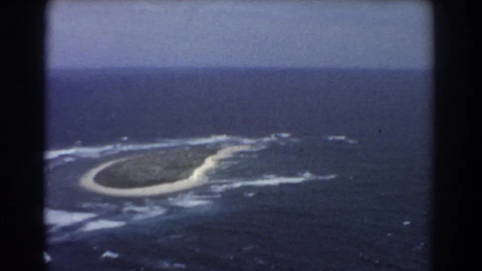 Vintage 8mm aerial frame near Perth, Australia, with a low island, bright surf line, and surrounding ocean under a hazy sky.