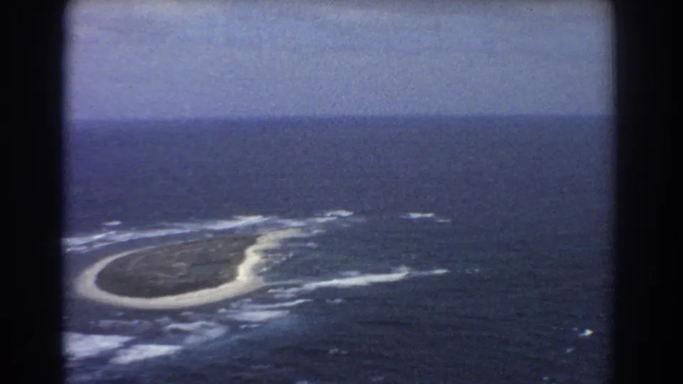 1967 home-movie aerial view near Perth showing the island receding in frame with surf, sand, and open sea visible.