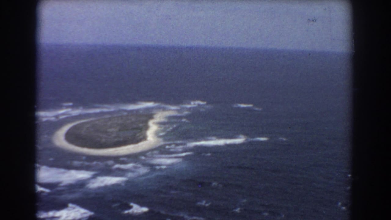 A small island seen from above, surrounded by dark ocean and white surf within black film borders.