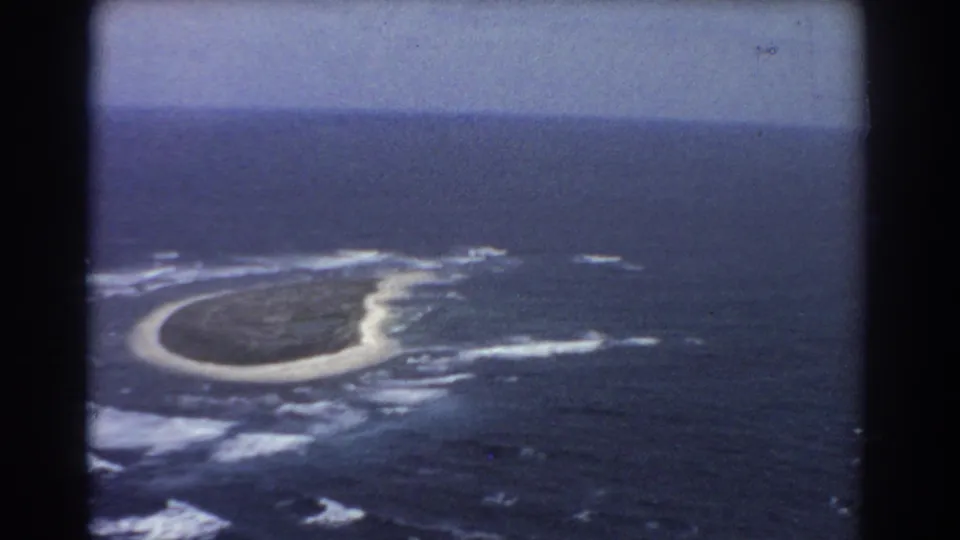 1967 8mm aerial view of a small island with pale shoreline, breaking surf, and deep blue water framed by dark projector borders.