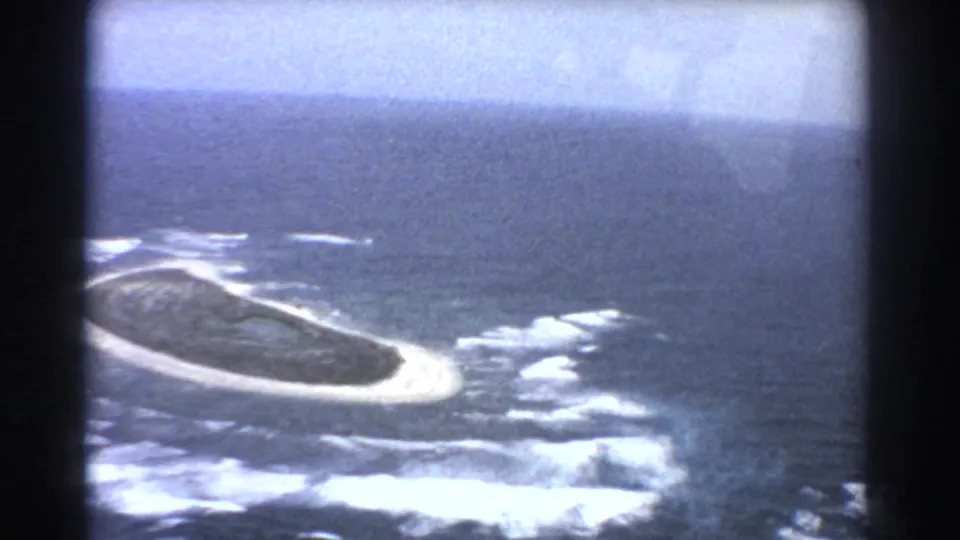 1967 8mm aerial view looking down toward a sandy island edge, white surf, reef-like shallows, and surrounding blue ocean.