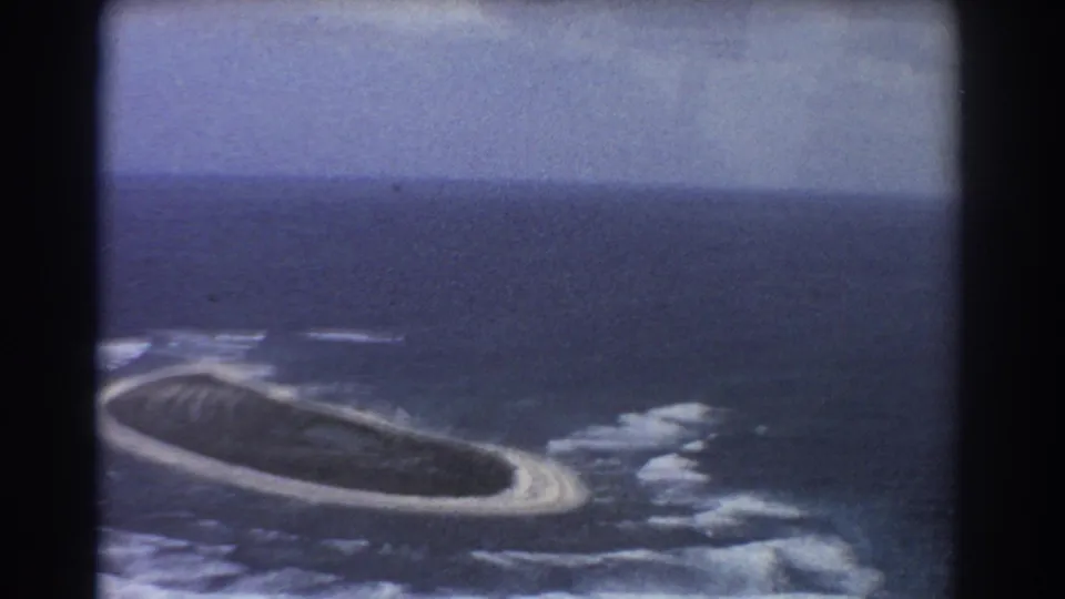 1967 8mm aerial view of an oval island, pale sand rim, breaking surf, open ocean, and hazy horizon in a soft home-movie frame.