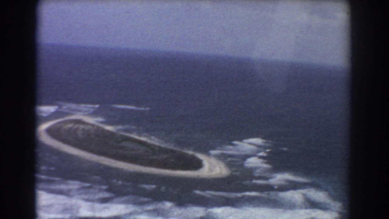 1967 Perth-area aerial 8mm view of a green oval island edged by pale sand and breaking waves.