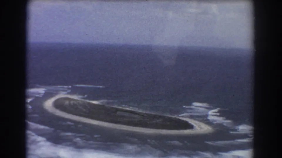 1967 8mm aerial pass over a green island near Perth, with surf lines and a pale beach perimeter visible.