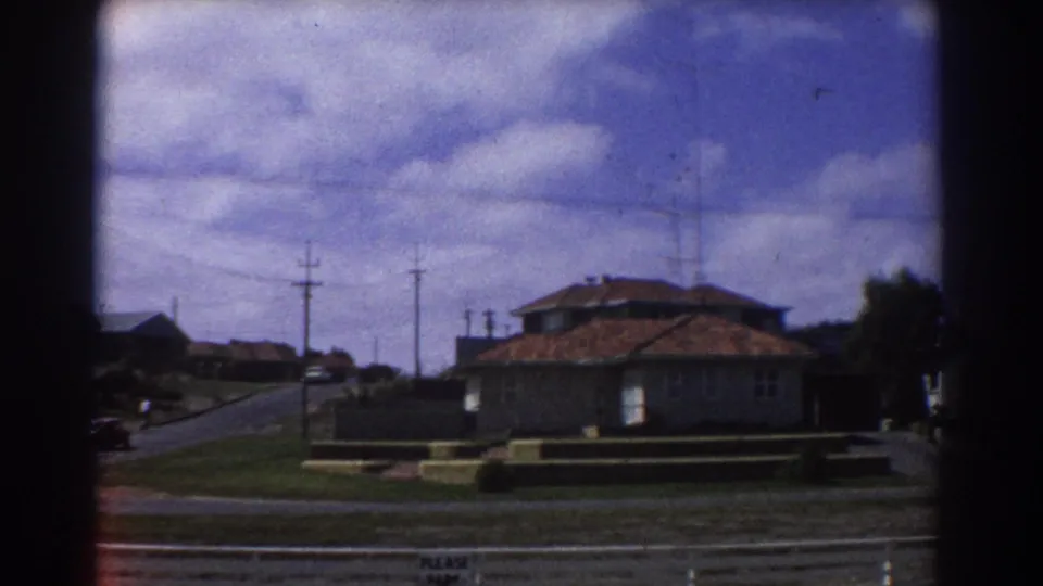 1967 Perth home-movie frame showing single-story suburban houses, red-tiled roofs, utility poles, and a partly cloudy sky.