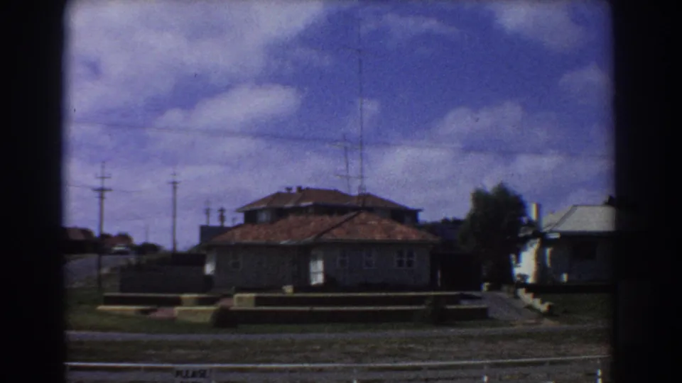 Perth, Australia, 1967 frame of a low house with a red-tiled roof, lawn, roadside edge, power poles, and clouded sky.
