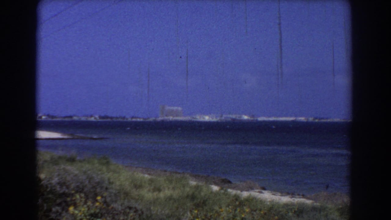 1967 Perth-area 8mm view of blue water, low grassy shore, and distant coastal buildings under open sky.