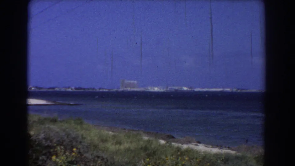 1967 Perth-area 8mm view of blue water, low grassy shore, and distant coastal buildings under open sky.