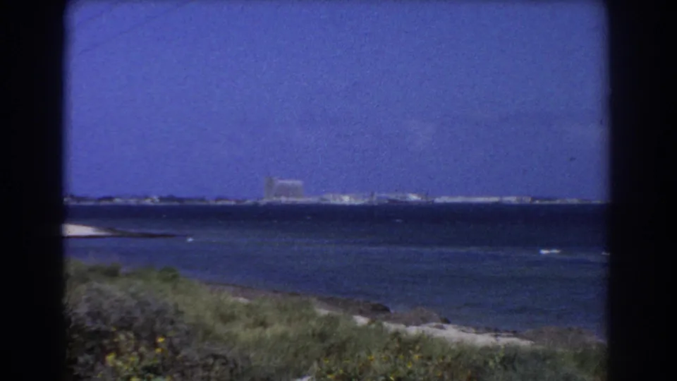 Perth-area 1967 coastal frame with blue water, grassy near shore, and a distant built-up strip on the horizon.