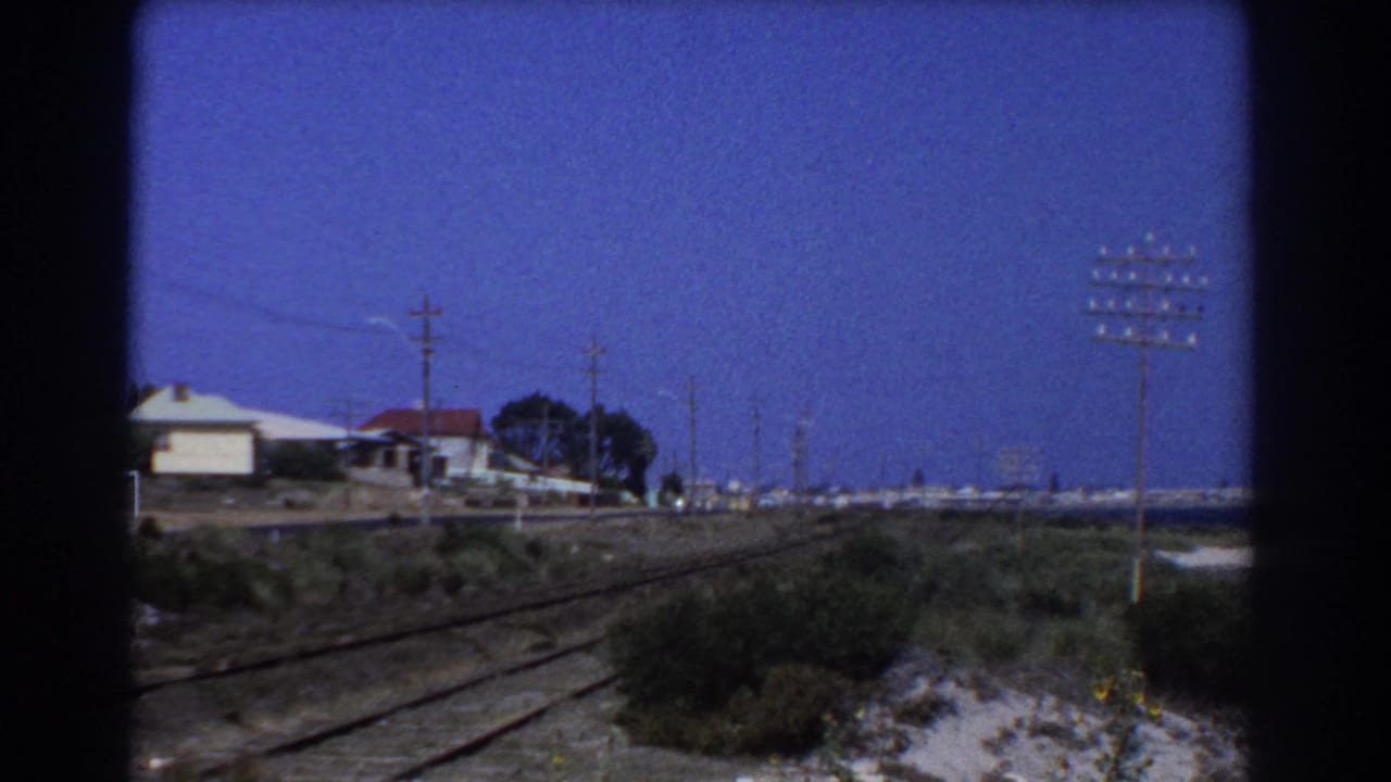 1967 Perth home-movie frame looking from a moving train toward rail tracks, low houses, and telephone or power poles.